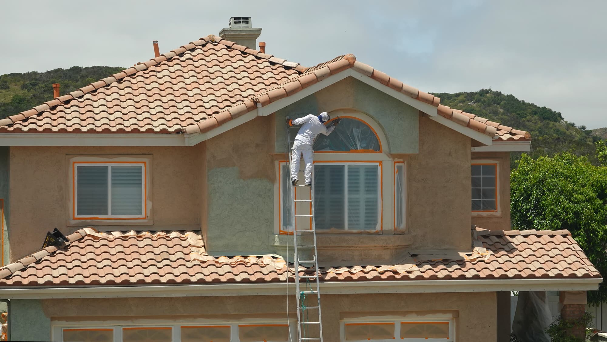 A painter on a ladder spraying the stucco of a two-storey tile-roofed home